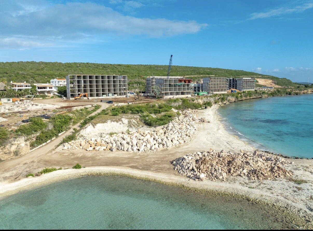 Aerial view of new hotel buildings under construction along a rocky Curaçao shoreline next to turquoise water, showing the development decisions shaping the island's growth